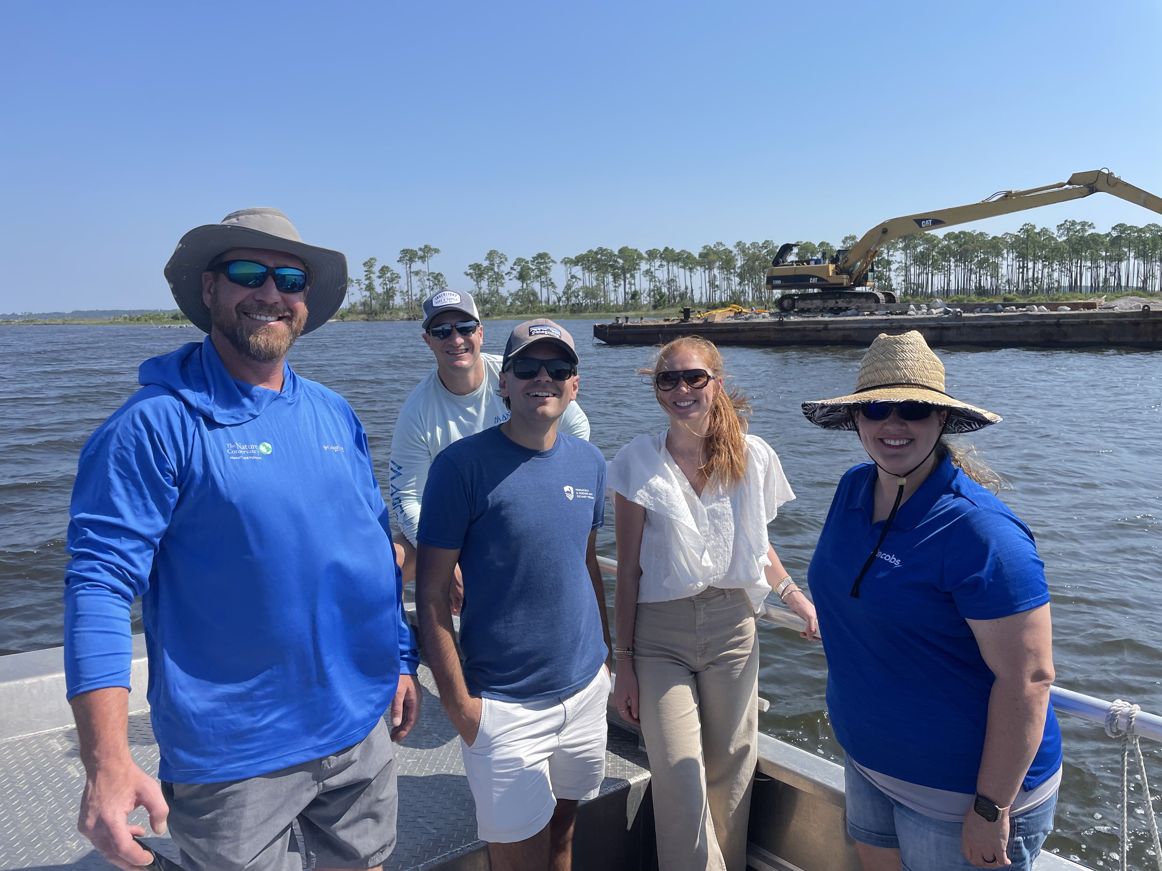 Five people on a boat in front of an oyster reef construction site with a barge and crane behind them